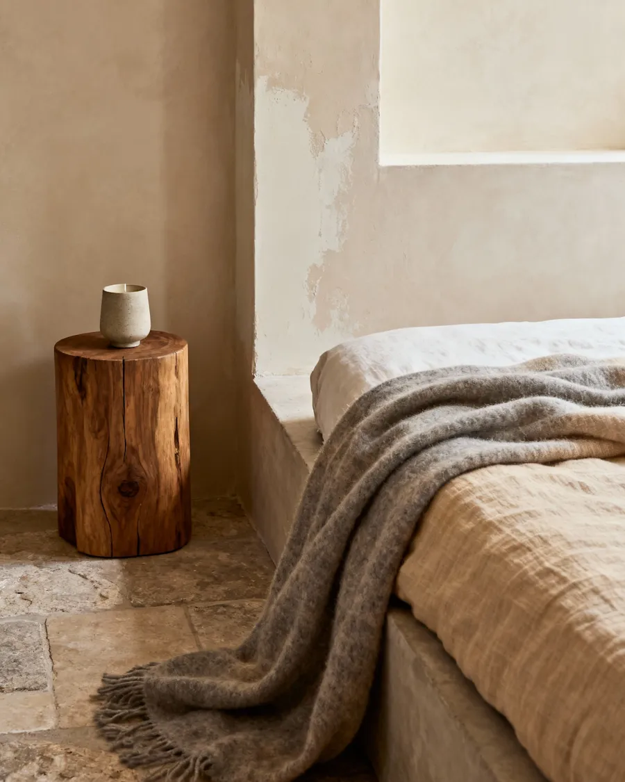 Bed with a grey wool throw beside a tree trunk side table on a stone floor