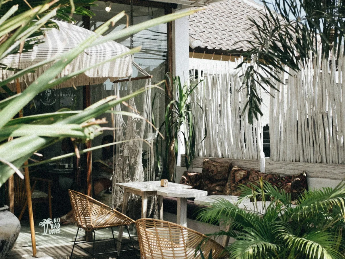 A shaded terrace with rattan chairs, white tables and large potted palms